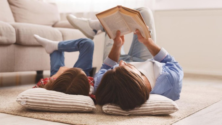 Little daughter and daddy enjoying time together, laying on floor at home, man reading book to his cute daughter, empty space
