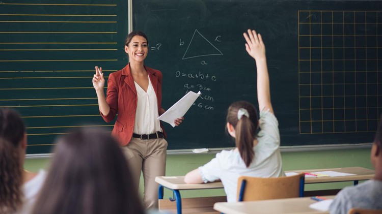 Teacher is standing in front of class and asking for volunteer. Pupils with hands raised are sitting in school desks and waiting for chance to show their knowledge in front of classmates. Back to school concept.