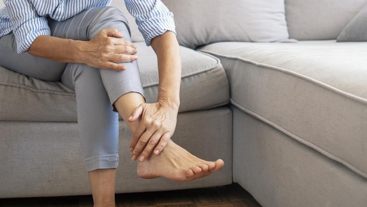 Closeup young woman feeling pain in her foot at home. Healthcare and medical concept. Tired and aching female feet after walking. Woman with feet intense pain sitting on a couch at home.