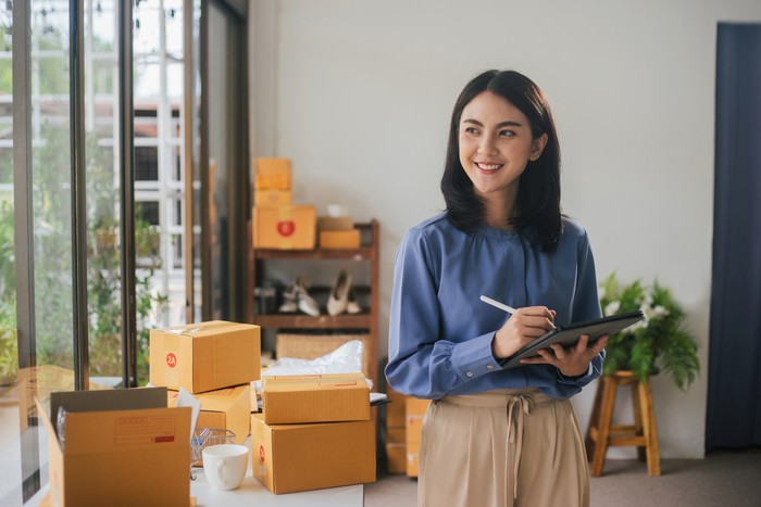 Portrait business Asian woman smile and use tablet checking information on parcel shipping box before send to customer. Entrepreneur small business working at home. SME business online marketing.