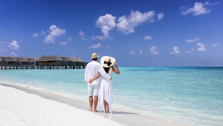 A hugging couple in white summer clothing stands on a tropical beach and enjoys the view to the turquoise sea and blue sky