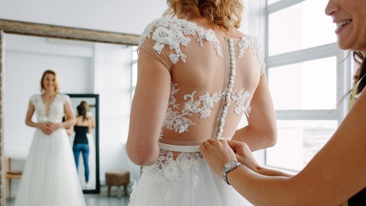 Bride is standing in front of mirror and fitting dress in atelier with wedding assistant. Woman with dressmaker are making final touch on tailor made gown in bridal clothing shop.