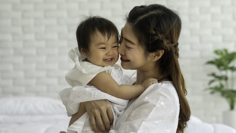 Happy Beauty mother Holding and kissing Cute Sweet Adorable Asian Baby wearing white dress sitting on Carpet smiling and playing with happiness emotional in cozy bedroom,Healthy Baby Concept