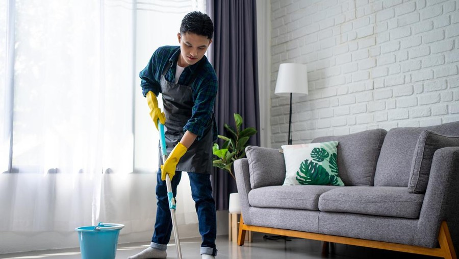 Handsome asian man wearing apron cleaning floor at home&period; Guy washing floor with mopping stick and bucket in living room&period;