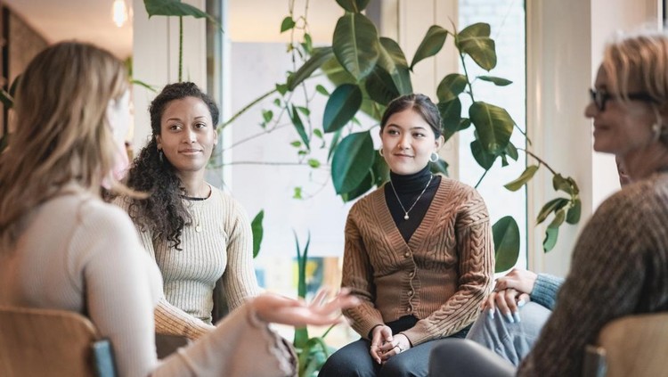1354857387 Woman sharing her experiences during a mental health group therapy meeting. Multiracial women participate in support group session sitting in a circle.
