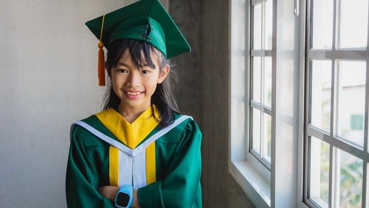 Portrait of cute Asian little girl wearing graduation hat and gowns standing in classroom at school, Elementary School and students concept