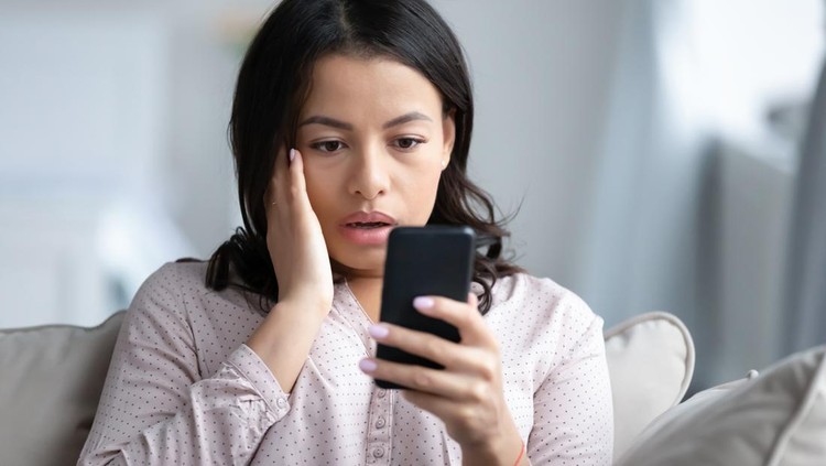 1208397282 Upset African American young woman sit on couch look at smartphone screen shocked by negative news online, sad stunned biracial female read bad unpleasant notice or message on cellphone gadget