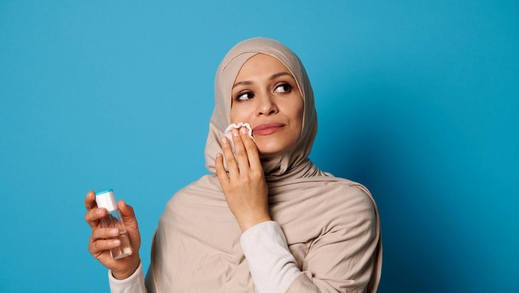 1311407763 Beautiful Muslim woman with covered head in hijab applying micellar lotion and removing make up from her lips. Make-up removal concepts on blue background with space for text