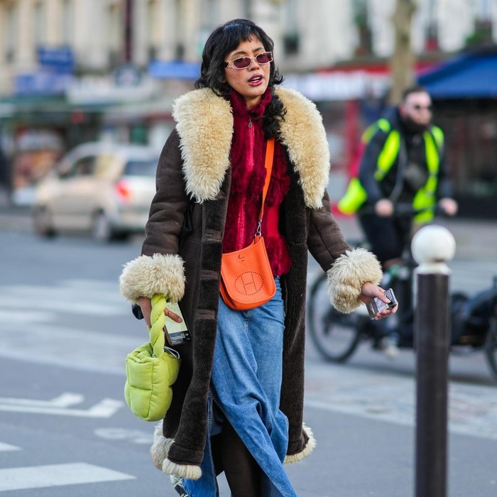 PARIS, FRANCE - JANUARY 18: A guest wears sunglasses from Chanel, a burgundy fluffy zipper high neck pullover, a brown shiny leather with large white latte sheep collar / wrist / borders long coat, an orange shiny leather Evelyne crossbody bag from Hermes, a blue denim jeans long slit / split skirt, a green quilted puffy handbag, black tights, rings, khaki / beige / black print pattern mules from Crocks, outside the Bluemarble show, during Paris Fashion Week - Menswear F/W 2022-2023, on January 18, 2022 in Paris, France. (Photo by Edward Berthelot/Getty Images)