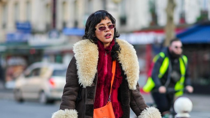 PARIS, FRANCE - JANUARY 18: A guest wears sunglasses from Chanel, a burgundy fluffy zipper high neck pullover, a brown shiny leather with large white latte sheep collar / wrist / borders long coat, an orange shiny leather Evelyne crossbody bag from Hermes, a blue denim jeans long slit / split skirt, a green quilted puffy handbag, black tights, rings, khaki / beige / black print pattern mules from Crocks, outside the Bluemarble show, during Paris Fashion Week - Menswear F/W 2022-2023, on January 18, 2022 in Paris, France. (Photo by Edward Berthelot/Getty Images)