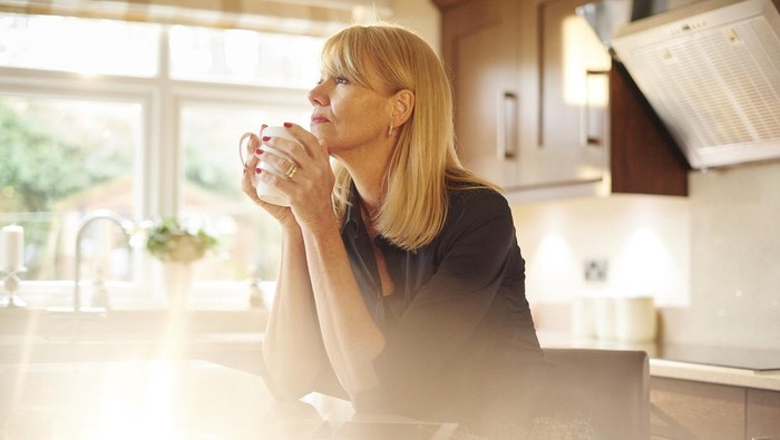 a woman sits in her kitchen and has a cuppa and a break looking off whilst she thinks.