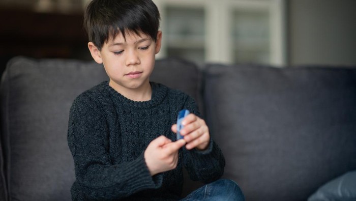 A boy is sitting in the living room. He is checking his blood sugar level because he is diabetic.