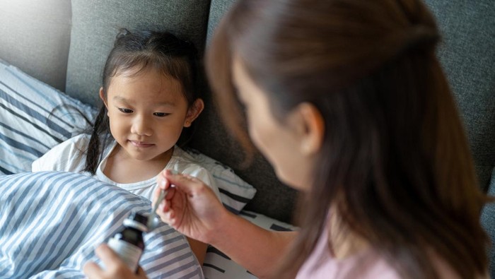 An Asian mom pouring cough syrup medicine into clear spoon to her daughter at bedroom,