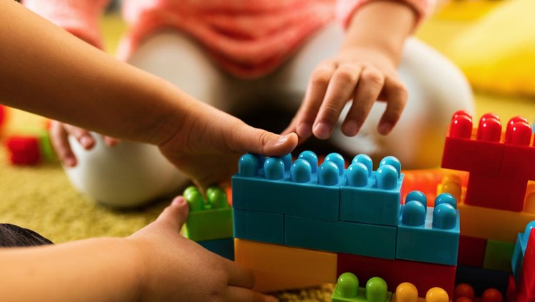 Ilustrasi Tempat Sewa Mainan Anak Close-up of two creative kids building something with plastic blocks on the floor.
