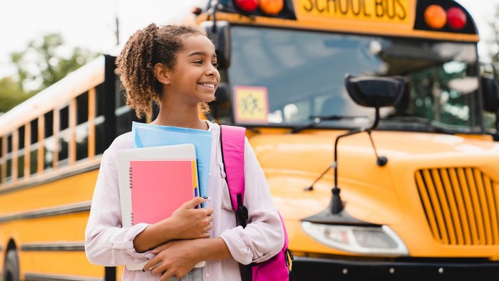 African-american girl teenager pupil student preparing to go to school after summer holidays holding books and notebooks standing next to the school bus.