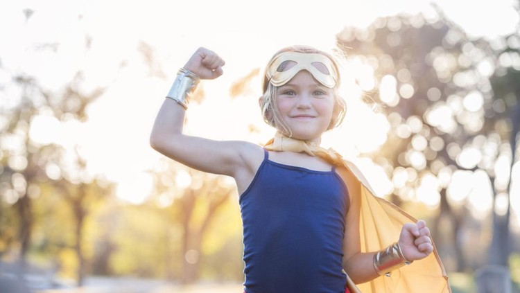Little girl enjoys dressing up as a superhero. She is smiling at the camera while flexing her muscles.