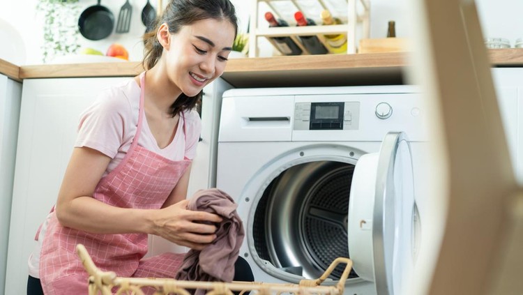 1350530979 Asian beautiful woman put dirty clothes to washing machine in house. Attractive girl wear apron sit on floor, feel happy to loads laundry in washer appliance at home. Domestic-House keeping concept.
