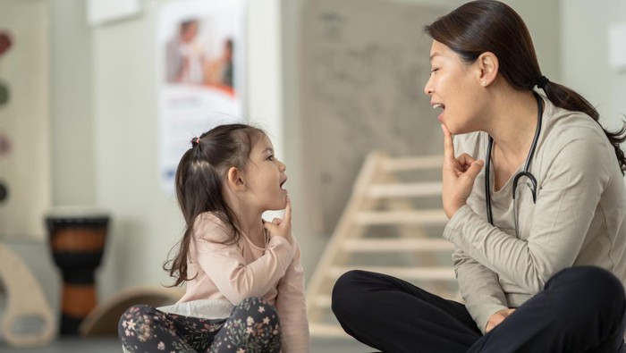 A female therapist sits beside her young patient on the floor as they work on some speech therapy exercises. They are each dressed casually and have their hands on their chins as they work through phonetical sounds.