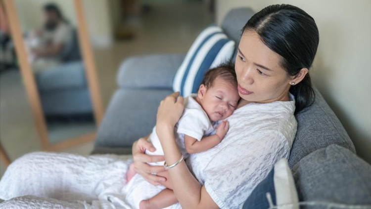 1347107958 Woman holding 40 days old newborn baby boy sitting on a sofa in a living room after feeding to help him burp at home. Mother hugging baby boy at home