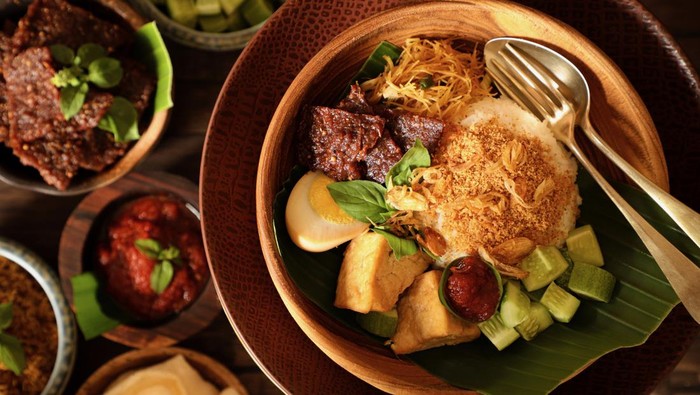 Nasi Ulam Betawi, a rice dish meal from Betawi cuisine in Jakarta. It consists of steamed rice topped with roasted grated coconut (Serundeng) and accompanied with beef jerky (Dendeng), soy-braised egg and beancurd (Semur Tahu Telur), fried rice noodles (Bihun Goreng), diced cucumber, basil leaves and red chili sauce (Sambal Oelek). The meal is plated in a shallow  wooden bowl lined with banana leaf. A set of spoon and fork is tucked in the bowl. The side dishes and condiments, each plated in its own serving dish, are arranged surrounding the rice dish.