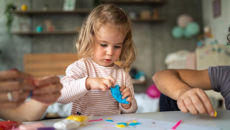 Ilustrasi Aktivitas Sederhana untuk Latih Motorik Halus Cute toddler girl making shapes of plasticine modeling clay, unrecognizable people next to her