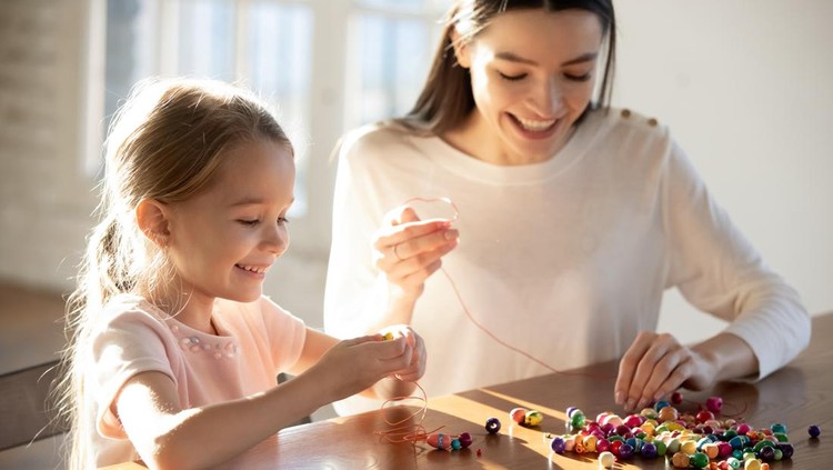 Happy caring young Caucasian mother and little 6s daughter have fun together making bracelets string colorful beads on thread. Smiling mom and small girl child involved in funny hobby activity.