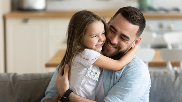 Close up smiling loving young father hugging adorable little daughter, enjoying tender moment, spending weekend together, sitting on cozy couch at home, good family relationship between dad and child