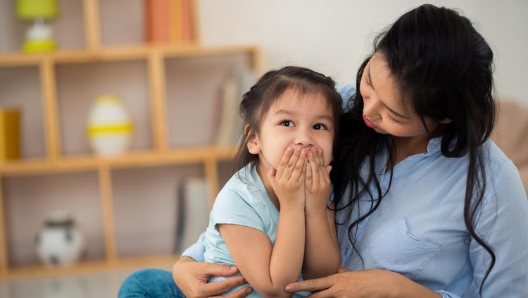 Ilustrasi Penyebab Anak Berbohong Portrait of surprised little girl sitting on laps of her pretty mother and covering mouth with hands, interior of cozy living room on background