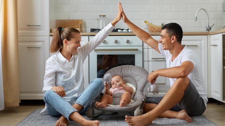 Full size photo of positive, three people mommy daddy small kid girl or boy in bouncer, parents make hands roof, enjoying sit on floor in light kitchen, indoors.