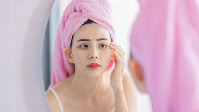 asian woman with towel on head looking mirror feel upset about dark rings under her eyes and touch her face