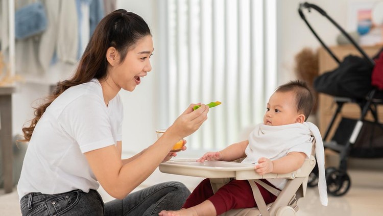 Side view mother feeding her asian baby daughter with pumpkin mashed or vegetable mash on rubber spoon.Mom trying to feed little baby boy at home enjoy and spending time together.Baby feeding Concept