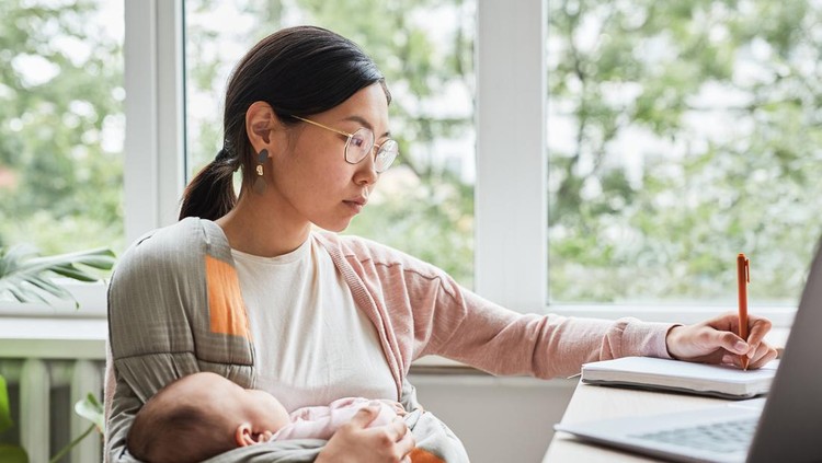 Asian young woman holding her baby on hands while making notes at the table she working at home