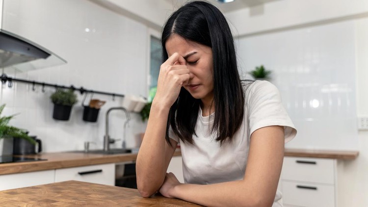 1351676669 Stressed young Asian woman with head in hand thinking. Exhausted female suffering from headache or migraine while sitting alone in the kitchen at home.