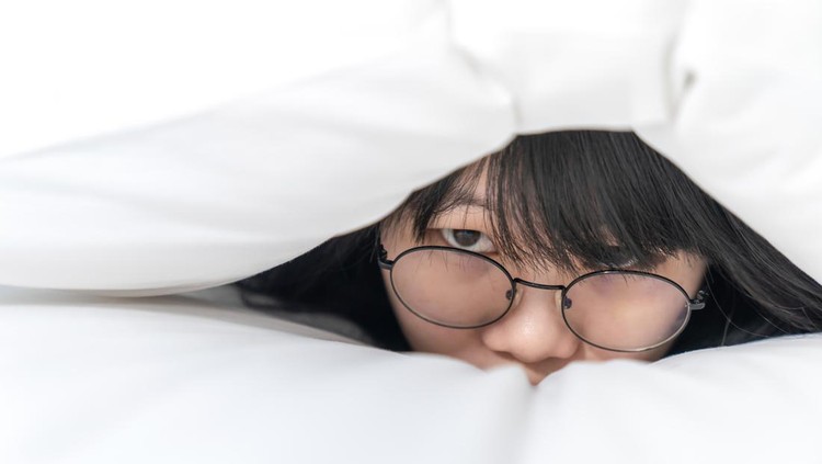 close up to the Cute Asian Glasses woman, she hides her body inside white pillow on the bed