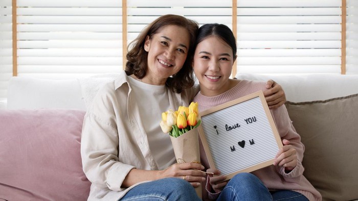 Attractive beautiful asian middle age mum sit with grown up daughter give gift box and flower in family moment celebrate mother day. Overjoy bonding cheerful kid embrace relationship with retired mom.