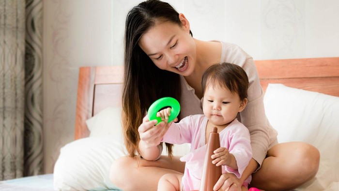 Asian and caucasian mom and child playing with colorful hoop toys on a bed at home. Mother teaching motor skills to her toddler.