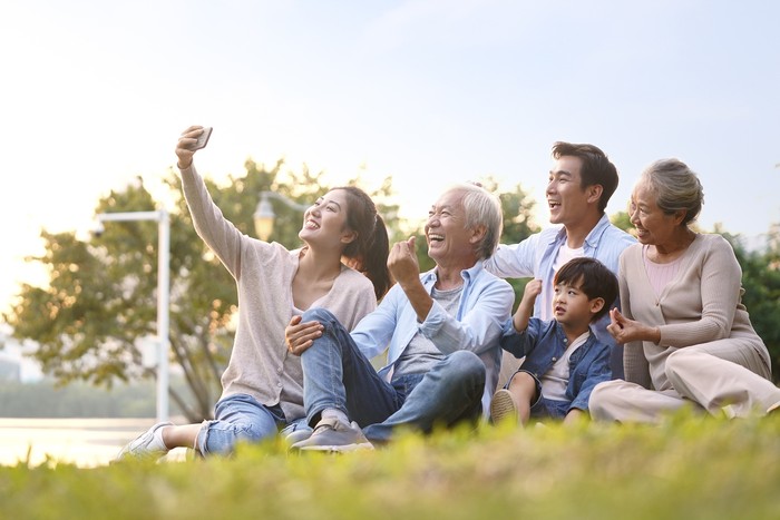 three generation happy asian family sitting on grass taking a selfie using mobile phone outdoors in park