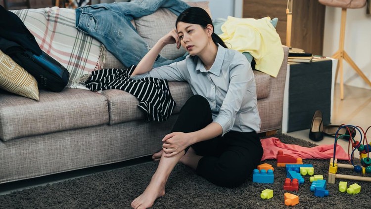 sleepy asian lady in blouse is sitting on floor with hand propping head on couch. japanese working mom taking rest in the parlor scattered with clothes and toys is feeling totally burnout.