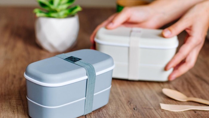 Two closed plastic two-layered lunch boxes with elastic band near wooden forks and spoons and paper cup on wooden table. Female hands holding one lunch box on the background. Selective focus on lunch box on the table.