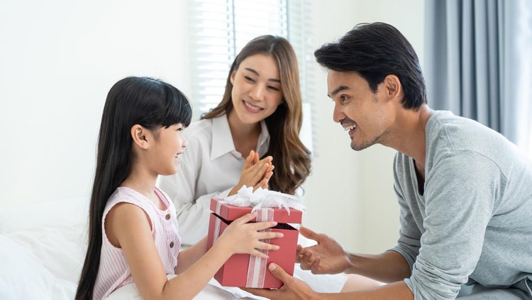 Asian lovely young little daughter open present gift box from parents. Happy family, kid feel happy and excited for surprise from mother and father ready to celebrate Birthday party together at home.
