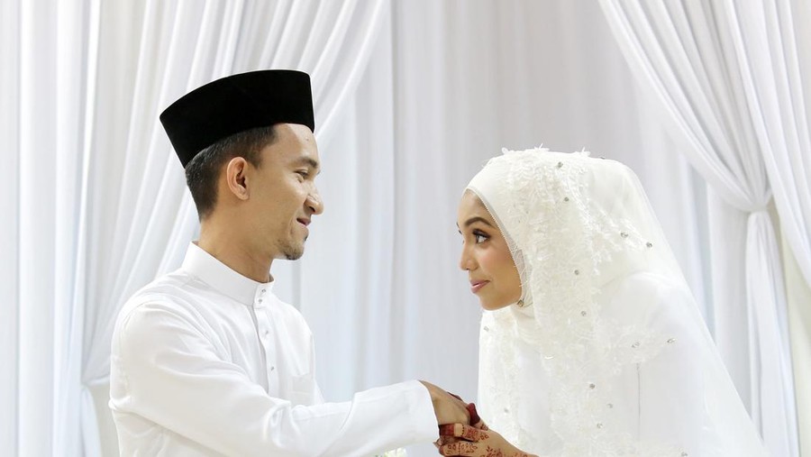 1211848386 A Muslim young woman holding a young man's hand for 'salam' (gesture of mannerism in greeting) when they are certified newlywed in Islam 'akad nikah' (solemnisation) ceremony. In this ceremony, newlywed and all guests are required to wear pure white tradiitonal clothing.