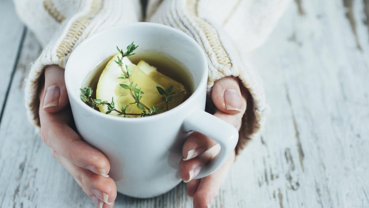 Human hands holding cup of tea with thyme herb and lemon slices on a wooden table,.