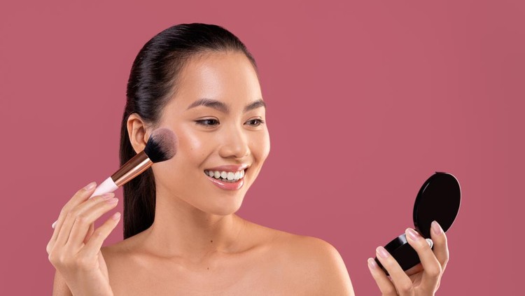 Pretty topless young chinese woman with makeup brush looking at mirror, applying blush on her cheeks, enjoying brand new makeup products on purple studio background, closeup photo
