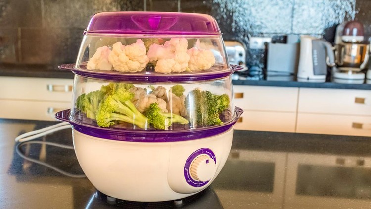 An electric two level plastic steamer loaded with cut vegetables on a kitchen island in a modern black and white kitchen interior