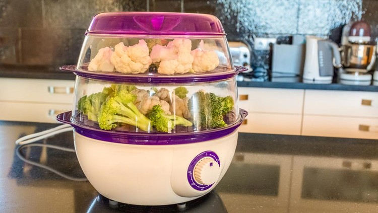 1047761826 An electric two level plastic steamer loaded with cut vegetables on a kitchen island in a modern black and white kitchen interior