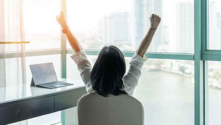 1216869584 Business achievement concept with happy businesswoman relaxing in office or hotel room, resting and raising fists with ambition looking forward to city building urban scene through glass window