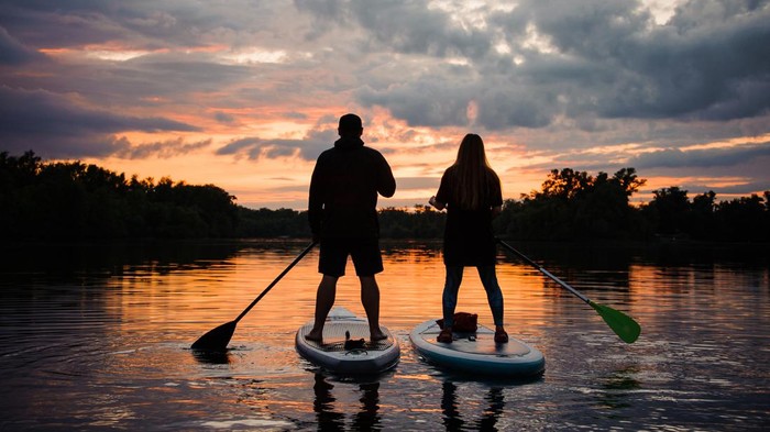 rear view on couple of people standing on sup boards with oars in their hands on the river at sunset