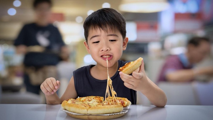 Asian 6-7 year boy is happy to eating pizza with a hot cheese melt stretched on a wooden pad in restaurant.