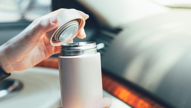 Close up of woman's hands holding reusable insulated water bottle and opening the lid in car in bright sunlight
