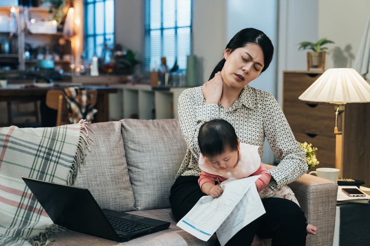 portrait asian working mother holding baby is having tension in her neck. millennial mom spraining her neck while looking after her child. lovely toddler is looking at document in her mother’s hand